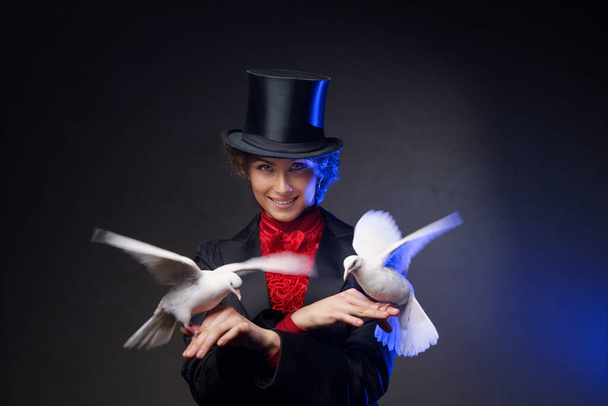 Eine fröhliche Zauberin in einem magischen Kostüm und schwarzem Zylinder führt zauberhafte Kunststücke mit weißen Tauben vor dunklem Hintergrund auf, der von blauem Licht erhellt wird - Foto, Bild