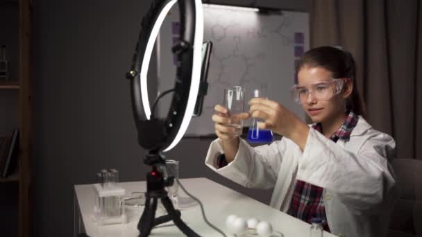 Carina blogger ragazza che mostra i suoi seguaci provetta con sostanza blu, mentre facendo esperimento di chimica in laboratorio a casa. Copia spazio - Filmati, video