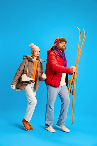 Full length portrait of father and daughter dressed winter outfit, warm suit and holds vintage ski and skates isolated blue studio background. Concept of winter, extreme sport, trip, parenthood. - Photo, image