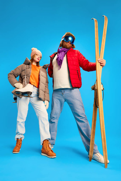 Funny portrait of father and daughter dressed winter outfit, warm suit and goggles mask and holds vintage ski and skates isolated blue studio background. Concept of winter, trip, relax, parenthood. - Photo, Image