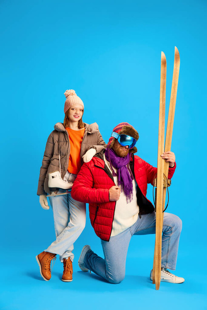 Father and daughter dressed winter warm outfit holds vintage ski and skates and posing looking at camera isolated blue studio background. Concept of winter, extreme sport, trip, relax, parenthood. - Photo, Image