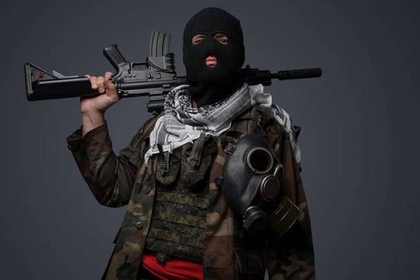Retrato amenazante de un soldado fanático radical de Oriente Medio vestido con un pasamontañas negro y un uniforme de campo camuflado, armado con un rifle automático, sobre un fondo gris frío - Foto, imagen