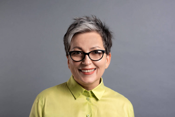 usmívající se 50s starší žena ve stylových brýlích s krátkými šedými vlasy při pohledu na kameru na šedém pozadí - Fotografie, Obrázek