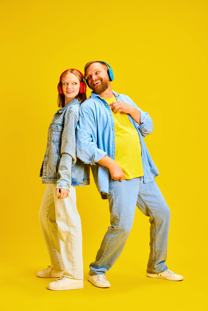 Батько і дочка, батько і дитина одягнені в джинсове вбрання і весело слухають музику в навушниках ізольовані жовтий студійний фон. Концепція батьківства, дитинства, сім'ї, стилю, моди. - Фото, зображення