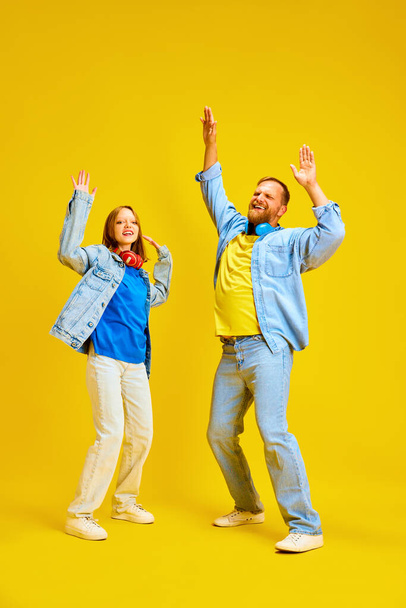 Szczęśliwy ojciec i córka, rodzic i nastolatek ubrane casual strój dżinsowy i zabawy, taniec podnosząc ręce w górę odizolowane żółte tło. Pojęcie rodzicielstwa, dzieciństwa, rodziny, stylu, mody. - Zdjęcie, obraz