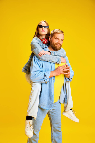Стильный джинсовый костюм для отца и дочери. Папа подвозит подростка на спине, ребенок сидит изолированный на ярком жёлтом фоне студии. Парфюмерия, детство, семья, стиль - Фото, изображение