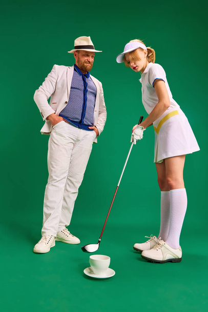 Vader en dochter hebben plezier op de golfbaan. Vader in slimme casual outfit met kopje koffie en gekleed als golf speler meisje tiener op groene achtergrond. Begrip ouderschap, hobby, recreatie. - Foto, afbeelding
