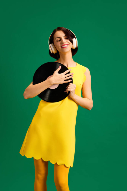 Portrait of young attractive woman dressed style yellow outfit dancing with retro plate while listening music in headphones isolated green background. Concept of youth, fashion, style, human emotions. - Photo, Image
