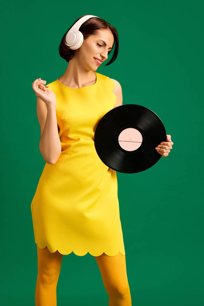 Half-length portrait of young woman dressed style yellow outfit dancing with retro plate while listening music in headphones isolated green background. Concept of youth, fashion, style, emotions. Ad - Photo, Image