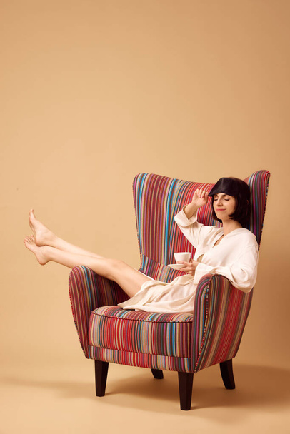 Jonge mooie dame zit in een fauteuil in zijden badjas en masker van slapen met een kopje hete koffie of thee geïsoleerde beige studio achtergrond. Concept van thuis levensstijl, ontspannen, wellness, zelfzorg. Steun - Foto, afbeelding