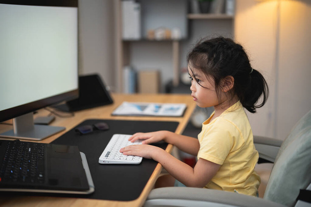 Azjatycka dziewczynka nosi t-shirt koncentrat do korzystania z laptopa i uczyć się online na drewnianym stole biurko w salonie w domu. Edukacja uczenie się online z koncepcji domu. - Zdjęcie, obraz