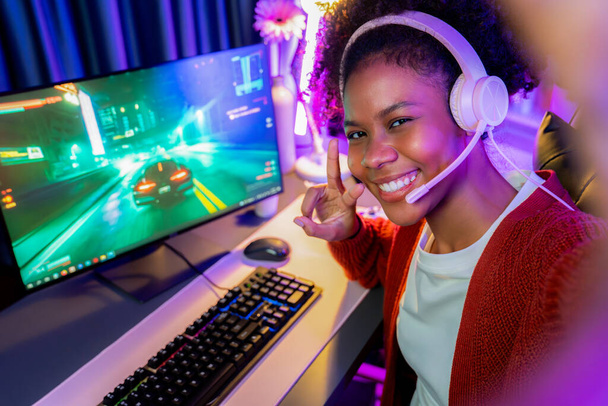 Host channel African of gaming streamer, African girl playing car racing game with joystick, wearing pastel headsets with mic, looking at the camera against background in neon color room. Tastemaker. - Zdjęcie, obraz