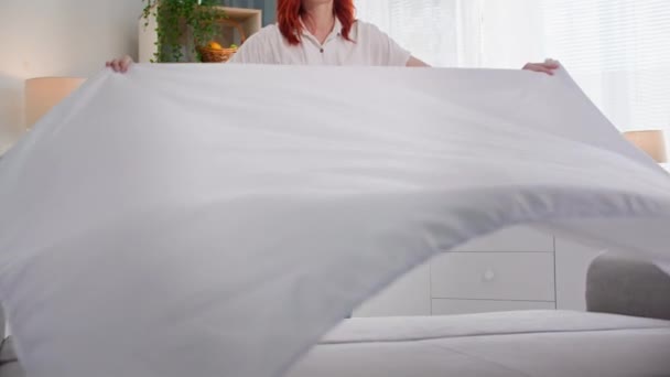 Güzel, genç bir ev kadınının portresi temizlik odası ve beyaz bir çarşafla yatak yapmak, gülümsemek ve kameraya bakmak. - Video, Çekim