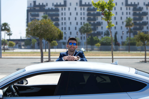 Joven hombre de negocios guapo con barba y traje azul apoyado en su coche deportivo blanco. El hombre lleva gafas de sol. Concepto de viaje empresarial - Foto, Imagen