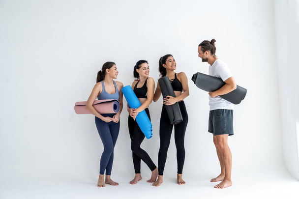 pełna długość uśmiechniętych młodych kobiet stojących z matami do jogi i rozmawiających na smartfonie stojąc przy ścianie w studio - Zdjęcie, obraz