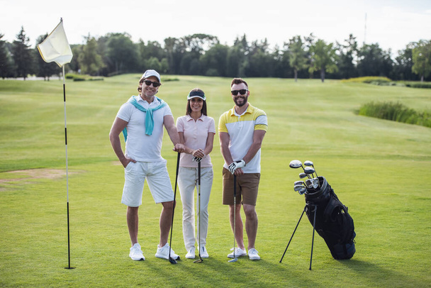 Dwóch mężczyzn i kobieta są gospodarstwa kije golfowe, patrząc na kamery i uśmiechając się stojąc na pole golfowe - Zdjęcie, obraz