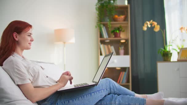 freelancer, charmante jonge vrouw werken op afstand met behulp van laptop computer terwijl liggend op de bank in de kamer - Video
