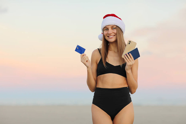 Sexy jonge vrouw in rode Kerstman hoed, bikini aan de kust houdt smartphone, creditcard en paspoort voor het boeken van online ticket om te reizen op Kerstmis en Nieuwjaar vakantie in tropische resort. - Foto, afbeelding