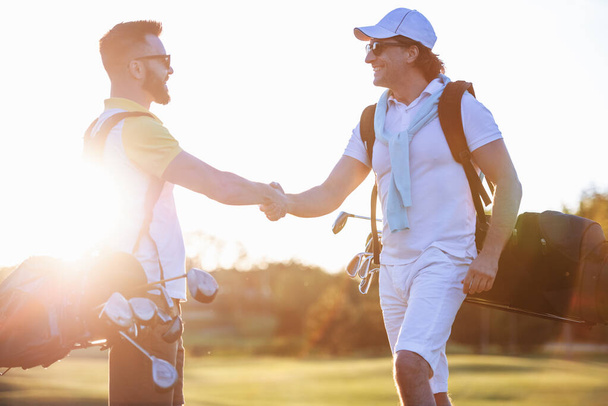 Homens bonitos estão apertando as mãos e sorrindo quando se encontram em um campo de golfe
 - Foto, Imagem