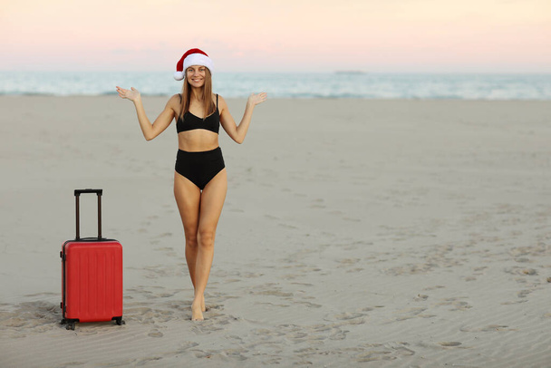 Vacaciones de invierno. Feliz joven sexy mujer en rojo Navidad Santa Claus sombrero, traje de baño con maleta en la orilla del mar muestra las manos para la publicidad reserva de entradas en línea, tours calientes para viajes vacaciones de Año Nuevo - Foto, Imagen