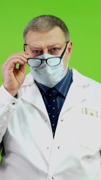 A fáradt főorvos leveszi a szemüvegét, maszkot vesz, és szalvétával törli meg a homlokát. Érett orvos a munkanap után, fáradt és pihenni akar. Zöld vászon, krómkulcs. Függőleges. - Felvétel, videó