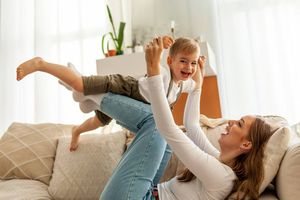 молода мати тримає і піднімає свого маленького сина на дивані, жінка грає зі своєю дитиною вдома, 2-річний хлопчик летить на руках свого батька - Фото, зображення