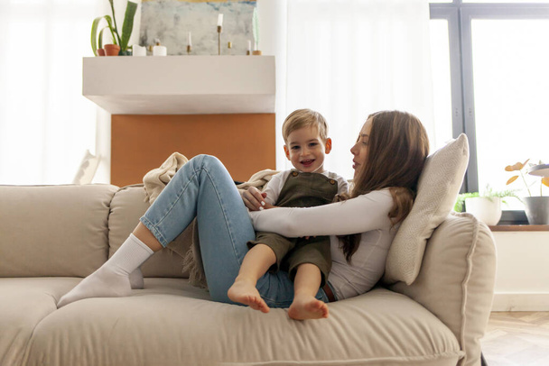 η νεαρή μητέρα με το γιο της κάθεται στον καναπέ και χαμογελά, η γυναίκα αγκαλιάζει το παιδί της στο σπίτι, το δίχρονο αγόρι αναπαύεται με τους γονείς του - Φωτογραφία, εικόνα