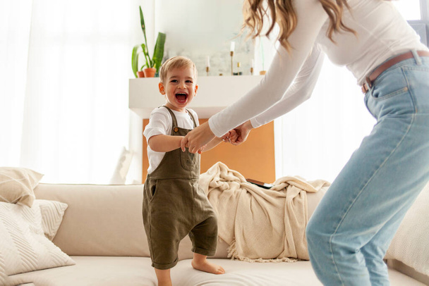 fiatal anya táncol és ugrál fiával a kanapén, nő játszik gyermekével otthon, 2 éves fiú örül és kiabál a szüleivel - Fotó, kép