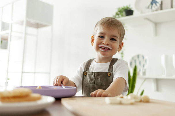 funny little boy, stained with food, eats yogurt and smiles in the morning in the kitchen, portrait of a 2-year-old child at the table having breakfast - Photo, Image