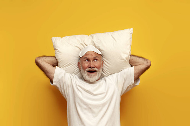 oude kale grootvader in pyjama en slaapmasker ligt op kussen en verbeeldt zich op gele geïsoleerde achtergrond, verrast oude man gepensioneerde dromen en kijkt - Foto, afbeelding