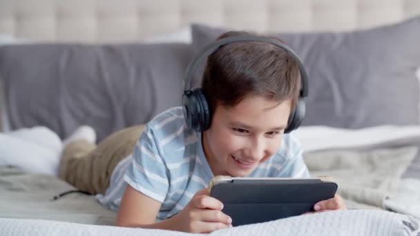 Ein kleiner Junge liegt bäuchlings auf dem Sofa im Schlafzimmer, benutzt ein Tablet und Kopfhörer. Ein Kind spielt ein Spiel, macht Grimassen und murrt mit sarkastischem zufriedenem Lächeln über den Sieg über das Internet - Filmmaterial, Video