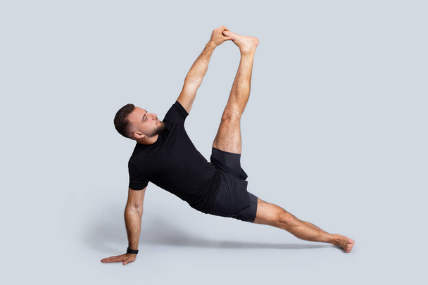 Poważny mężczyzna rasy kaukaskiej w sportowej jodze treningowej, rozciągający nogi, ćwiczący oddech, cieszący się sportem, odizolowany na szarym tle studia. Zdrowie i pielęgnacja ciała, styl życia i witalność - Zdjęcie, obraz
