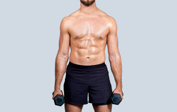 Потный мускулистый молодой европейский спортсмен с обнаженным туловищем, делает упражнения с гантелями, изолированные на сером студийном фоне, обрезанные. Сила, силовые тренировки для мышечных бицепсов - Фото, изображение