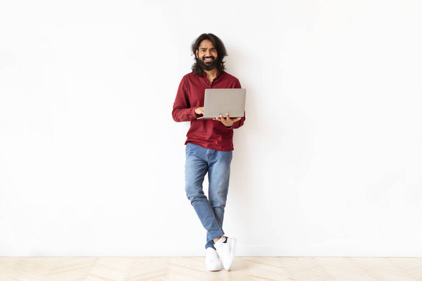 Pozitivní pohledný vousatý tisíciletý indián chlap na volné noze nezávislý dodavatel drží počítač notebook, pózovat na bílém pozadí, kopírovat prostor. Online vzdělávání, vzdálená práce - Fotografie, Obrázek