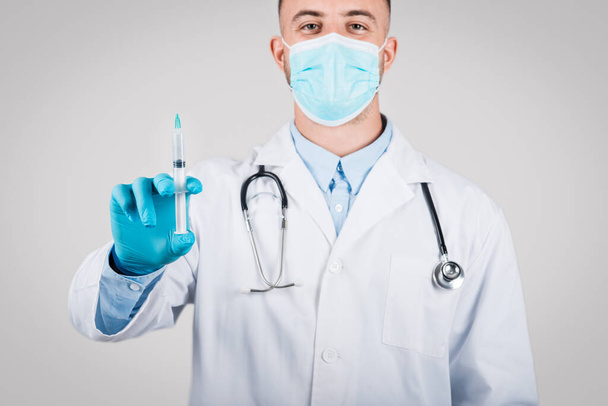 Professionnel de santé masculin avec stéthoscope et masque de protection présentant une seringue médicale, prêt à être vacciné, debout sur fond gris - Photo, image