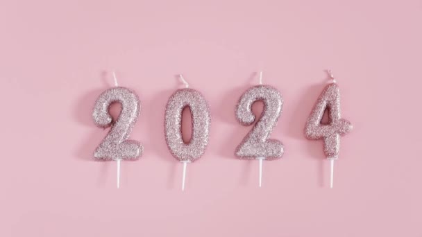 Een tak van gouden kerstbladeren, verspreid confetti, snoepjes en feestelijke glanzende kaarsen nummer 2024 liggen op een roze achtergrond, plat van dicht bij elkaar met een cirkelvormige rotatie naar links. - Video