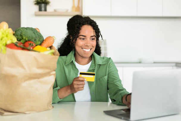 Jeune femme noire avec des sourires de carte de crédit comme elle navigue sur le site d'épicerie sur ordinateur portable assis à la table de cuisine moderne à la maison, faire du shopping des produits alimentaires et websurfing recettes saines - Photo, image