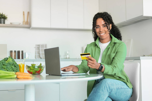 アフリカ系アメリカ人女性が,自宅のキッチンでクレジットカードとラップトップでオンラインで食料品を注文し,テーブルに座って笑顔でカメラに向かう. 金融に精通した現代的なライフスタイル - 写真・画像