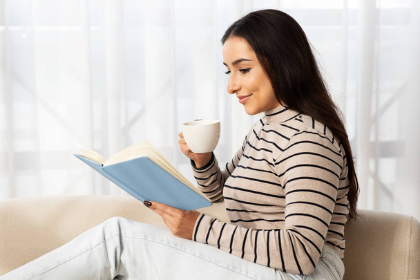 Ruhige junge hübsche Araberin liest Buch, plant Arbeiten, sitzt auf dem Sofa, trinkt Kaffeetasse im hellen Wohnzimmer. Lady genießt Freizeit, Studium, Bildung, Hobby und Lifestyle gemütlich zu Hause - Foto, Bild