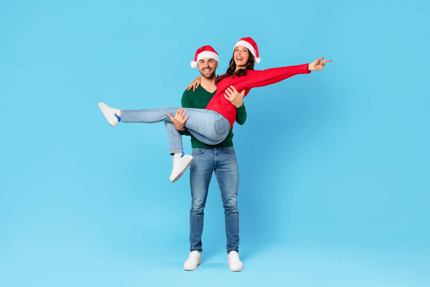 Joyful european couple in Santa hats having fun while posing in studio, man carrying woman celebrating Christmas and New Year holidays over blue backdrop. Full length, copy space - Photo, Image