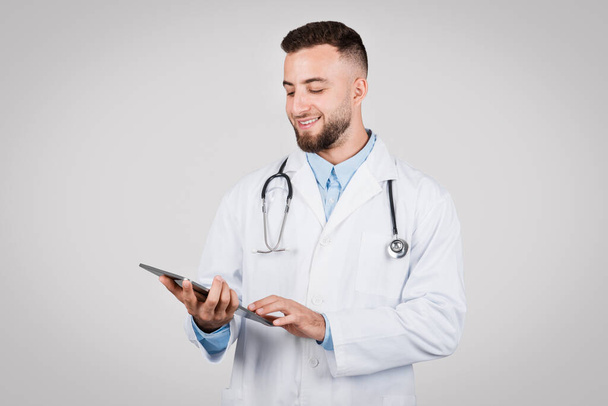 Médico masculino comprometido con bata de laboratorio blanca mirando la tableta digital, indicativo de las prácticas sanitarias modernas y la telemedicina, contra el fondo gris - Foto, imagen