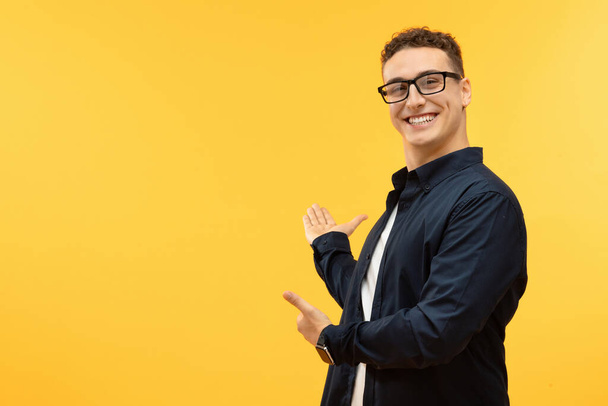 Positive handsome young man wearing casual outfit and eyeglasses showing blank space and smiling, isolated on yellow background. Blank space for text, advertisement - Photo, Image
