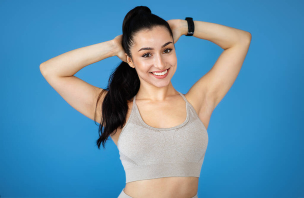 Sports lifestyle, fitness, gym concept. Portrait of happy sporty attractive young woman with hands behind head in beige sportswear smiling at camera on blue studio background - Photo, Image