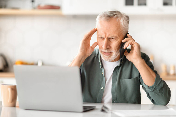 Stressed senior man using phone and laptop while sitting at desk in kitchen, worried elderly male having problems with remote work, suffering troubles with multitasking at home, copy space - Photo, Image