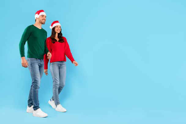 Weihnachtsangebot. Glückliches europäisches Paar in Weihnachtsmützen und lässiger Kleidung, stehend auf blauem Hintergrund, mit Blick auf freien Platz für Neujahrs- oder Weihnachtswerbung, in voller Länge - Foto, Bild