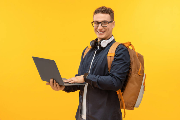 Счастливый красивый молодой человек в повседневной одежде, неся рюкзак студента с ноутбуком на желтом студийном фоне, наслаждаясь онлайн-классами. Концепция электронного образования - Фото, изображение