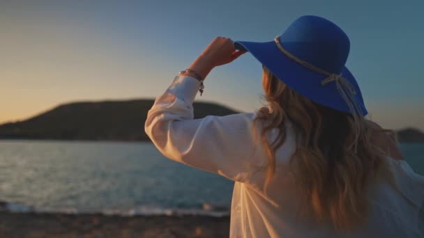 Gün batımında deniz kenarında mavi şapkalı bir kadın. - Video, Çekim