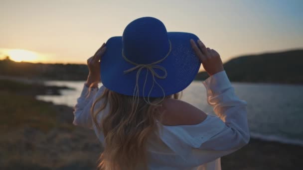 Gün batımında deniz kenarında mavi şapkalı bir kadın. - Video, Çekim