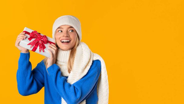 Fröhliche junge Frau in Winterkleidung hält aufgeregt ein Weihnachtsgeschenk in der Hand und zeigt Freude und Vorfreude vor leuchtend gelbem Hintergrund - Foto, Bild