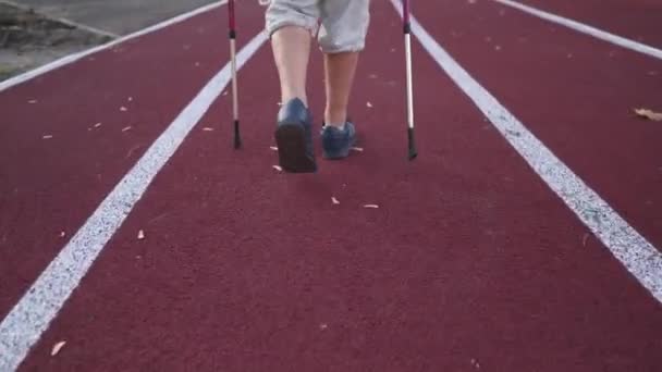 Primo piano di una donna anziana che cammina su una pista da stadio con pali da nordic walking vista posteriore. Stile di vita attivo in età avanzata e esercizi di resistenza cardio con bastoni sportivi. Passeggiata scandinava - Filmati, video